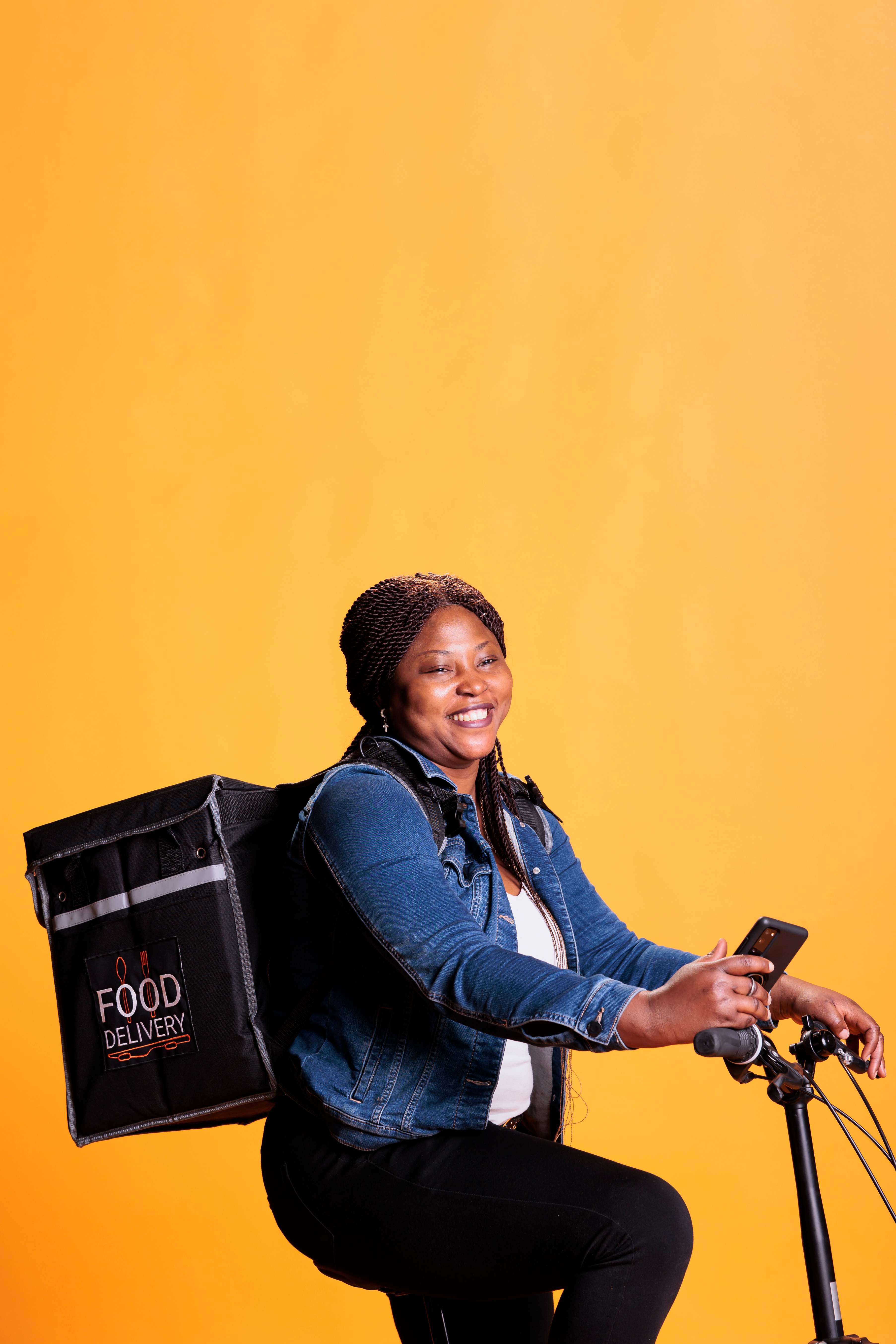African courier with delivery backpack and bicycle, smiling while using smartphone to check delivery address on yellow background